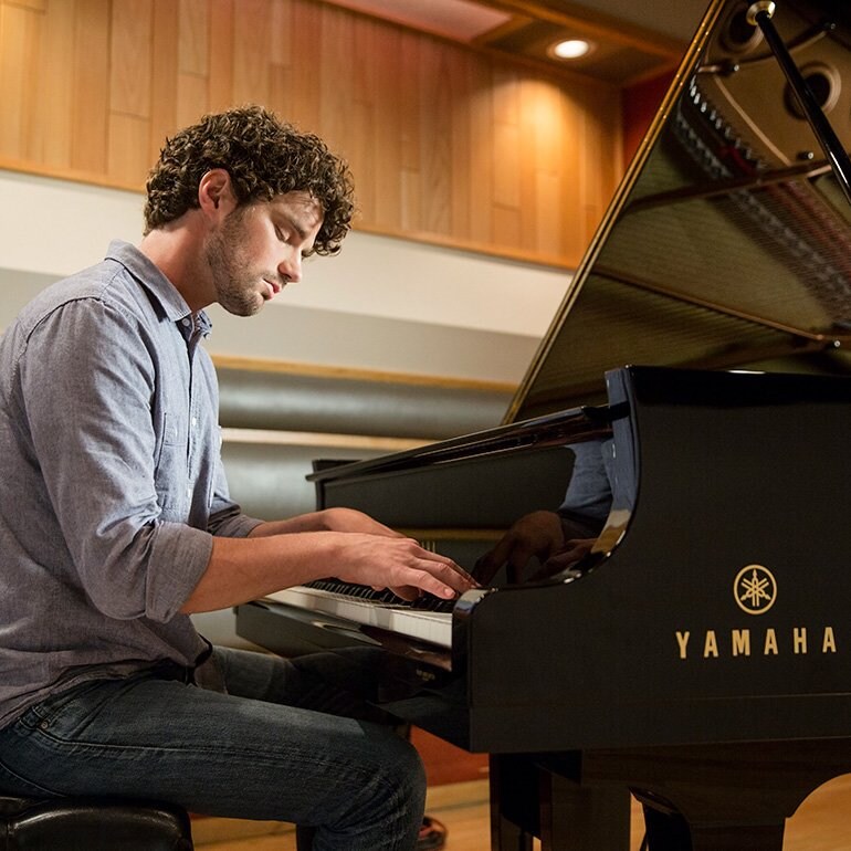A man playing a Yamaha grand piano in a wood-paneled studio.