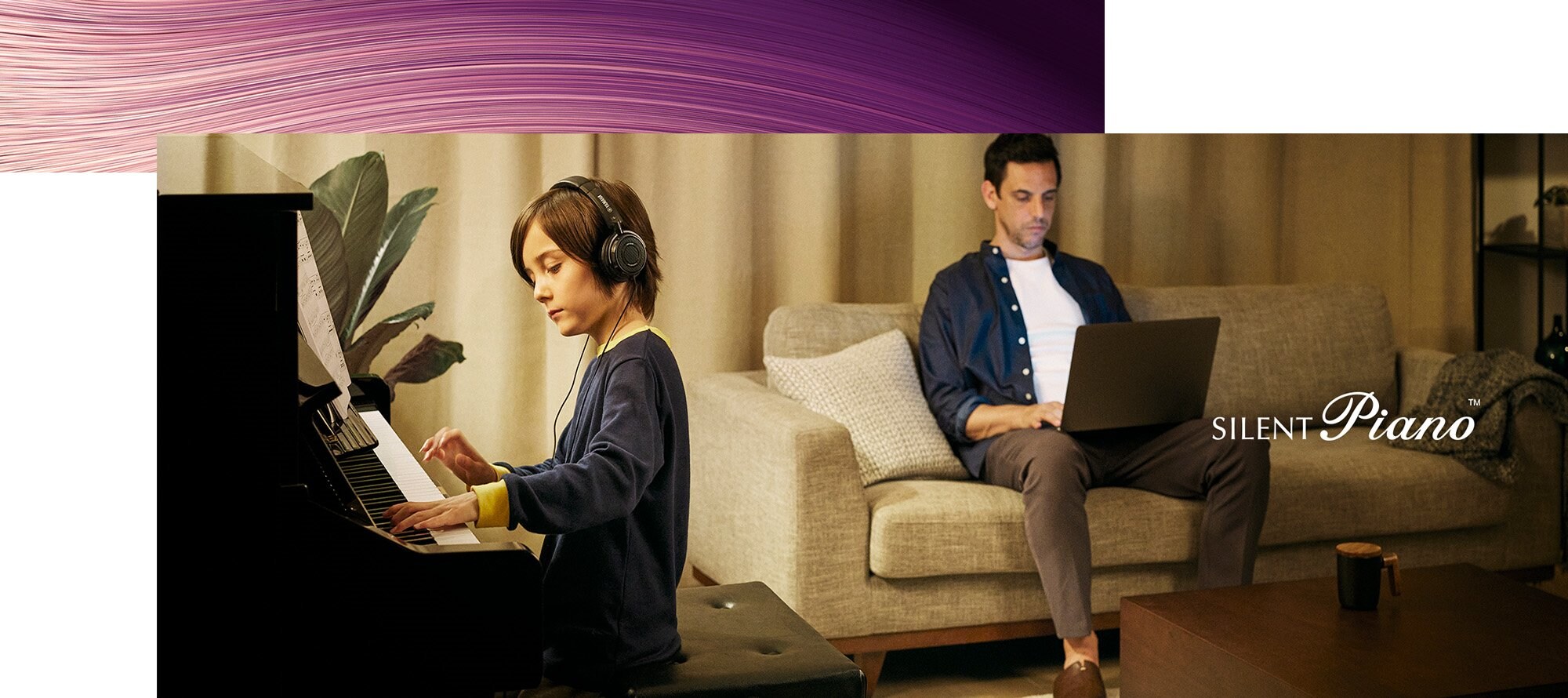 A young boy wearing headphones playing a Yamaha SILENT Piano while an adult works nearby.